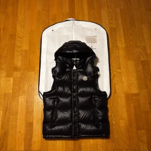 Moncler Bormes - Svart, glansig dunväst från Moncler med avtagbar huva och dragkedja framtill. Västen har två fickor med dragkedja och klassisk Moncler-logga på bröstet. Perfekt för lager-på-lager och riktigt snygg till streetwear. Storlek 3 motsvarar L/M. Ser helt nya ut nästan oanvänd, kvitto finns. Säljer pågrund av att jag sparar till bil och behöver snabba pengar. Vid snabb affär kan gå ner i pris. Om frågor kom pv!