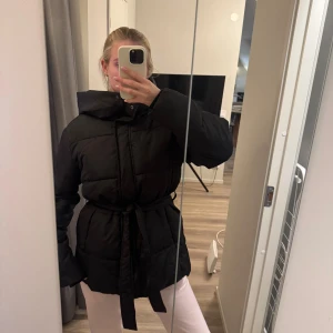 Svart pufferjacka med bälte och huva - Säljer en svart pufferjacka med bälte i midjan och stor huva. Jackan har långa ärmar, quiltad design och stängs med dragkedja och tryckknappar. Perfekt för kalla dagar och riktigt snygg till alla outfits. Storlek xs men är stor i storlek så skulle säga som en S