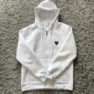 Comme des Garçons zip hoodie - Säljer en vit hoodie från Comme des Garçons Play i storlek M. Tröjan har dragkedja, huva med snörning och fickor framtill. Ikoniskt svart hjärta med ögon på bröstet och stort tryck på ryggen. Perfekt för dig som gillar streetwear och stilrena detaljer. Säljer då den inte passar mig.