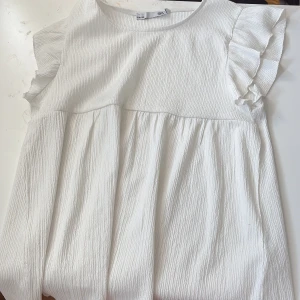 Vit kortblus med volangärmar från 157 - Supersöt vit kortblus från 157 med volangdetaljer på ärmarna och rund hals. Blusen har en lätt rynkad kjoldel och är tillverkad i ett strukturerat bomullstyg som ger en skön och luftig känsla. Perfekt för dig som gillar enkel och fräsch stil.