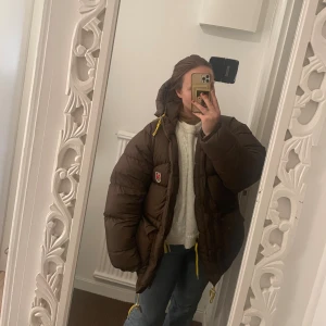 Brun pufferjacka från Fjällräven - Säljer en brun pufferjacka från Fjällräven med huva och gula snören. Jackan har stora fickor med knappar, dragkedja och Fjällräven-logga på bröstet. Är en äldre modell. 