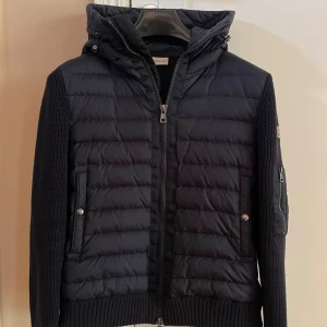 Svart dunjacka från Moncler med huva - Svart Moncler dunjacka med stickade ärmar och quiltad front. Jackan har huva, dragkedja framtill, fickor med tryckknappar och en extra ficka med dragkedja på ärmen. Klassisk Moncler-logga på vänster ärm. Perfekt för kyliga dagar.