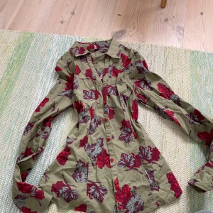 Grön blommig skjorta  - Snygg grön skjorta med röda och lila blommönster. Skjortan har lång ärm, klassisk krage och knappar framtill. Materialet känns lätt och skönt mot huden, perfekt för dig som gillar färgstarka prints, den är i storlek L men funkar för någon som har S eller medium också