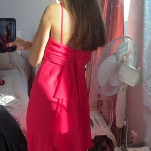 Lång rosa klänning med smala axelband - Säljer en elegant rosa långklänning med smala axelband och snygg drapering över bysten. Klänningen har en vacker detalj med rosett och veckad rygg, vilket ger en extra touch. Materialet känns lätt och flowy, perfekt för dig som vill sticka ut med en färgstark look.