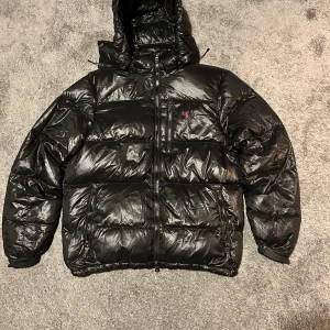 Svart pufferjacka Ralph Lauren M - Svart pufferjacka från Polo Ralph Lauren med glansig finish, röd broderad logga på bröstet och huva. Jackan har dragkedja framtill och två sidofickor med dragkedja. Perfekt för kalla dagar och riktigt snygg streetstil. Tyvär ett litet hål på baksidan och därav priset men jag kan laga det om köparen vill anars riktigt bra skick