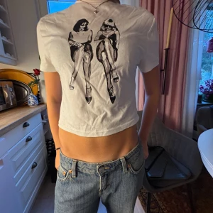 Vit croppad t-shirt med tryck - Säljer en vit croppad t-shirt med korta ärmar och ett svart tryck framtill som föreställer två sittande personer. T-shirten är i mjuk bomull och har en avslappnad passform. Perfekt för dig som gillar unika prints och vill sticka ut lite extra. 