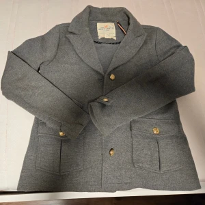 Grå kavaj från Holly & White - Stilren grå kavaj från Holly & White med guldknappar och två stora fickor framtill. Klassisk krage och långärmad modell i mjukt material som ger en dressad men ändå chill look.