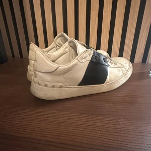 Valentino Open sneakers vit/svart box medkommer - Snygga Valentino Open sneakers i vitt skinn med bred svart läderdetalj över snörningen. Klassisk rund tå och platt sula med mönstrad undersida. Ikoniska nitar längs sidan av sulan ger extra edge. Perfekt för dig som gillar exklusiva streetwear-vibbar. 