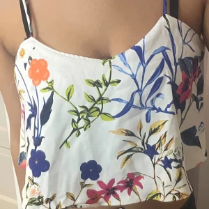 Vit blommig linnetopp med tunna axelband - Säljer en fräsch vit linnetopp med färgglatt blommigt mönster i blått, rosa, orange och grönt. Toppen har tunna axelband och är croppad med en luftig passform. Perfekt för varma dagar och ger en riktigt somrig vibe.