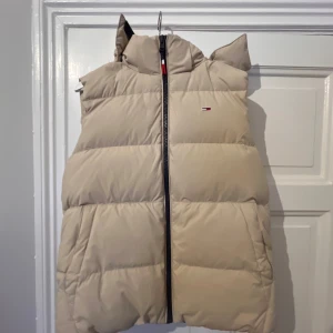 Beige dunväst från Tommy Hilfiger - Beige dunväst från Tommy Hilfiger med avtagbar huva och dragkedja framtill. Västen är quiltad och har en klassisk puffer-look med diskret logga på bröstet. Perfekt för lager-på-lager och snygg till både hoodie och t-shirt.