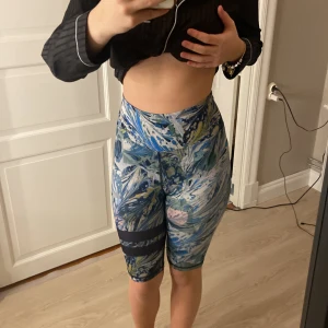 Färgglada träningstights shorts - Säljer ett par höga, tighta träningstights shorts med färgstarkt abstrakt mönster i blått, grönt, vitt och inslag av svart. De har bred resår i midjan och svarta ränder på ena benet. Perfekta för gymmet eller löprundan.