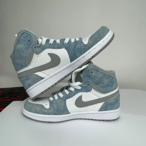 Nike Air Jordan 1 High OG blå/vit - Nike Air Jordan 1 High OG med blå mockadetaljer och vita paneler i skinn. Grå swoosh och häl, vita skosnören och sula. Klassisk high-top siluett med perforerad tåbox och blå Nike Air-logga på plösen. Perfekta för dig som vill sticka ut med stilrena sneakers.