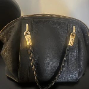 Svart handväska från Michael Kors - Snygg svart handväska från Michael Kors i äkta skinn med grov struktur. Väskan har guldfärgade metalldetaljer, flätat handtag och dragkedja upptill. Insidan är fodrad med logotypmönster och har flera fack för dina grejer.