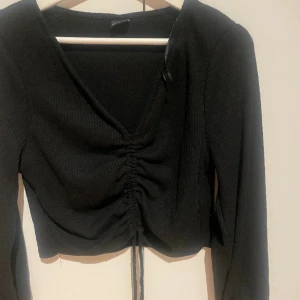 Svart ribbad croppad topp med snörning - Säljer en svart långärmad topp med ribbad struktur och v-ringning. Toppen är croppad och har snygg snörning framtill som ger en cool rynkad effekt. Perfekt för dig som gillar stilrena och trendiga plagg med lite edge.