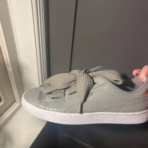 Grå Puma Basket sneakers med breda snören - Snygga grå Puma Basket sneakers med breda, ribbade snören och vit platt sula. Skorna har en blank finish i syntetmaterial och guldfärgad Puma-logga på sidan. Oanvända av mig men köpta på sellpy🩶