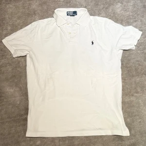  Ralph Lauren pike - Klassisk vit pikétröja från Polo Ralph Lauren med korta ärmar, krage och knappar framtill. Tröjan har den ikoniska broderade loggan på bröstet och är tillverkad i mjuk bomull. Perfekt för en clean och stilren look. Storlek S