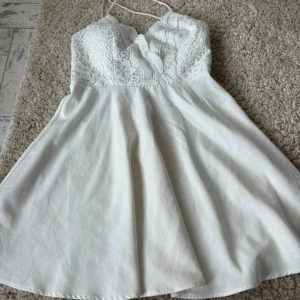 Studentklänning  - Supersöt vit kortklänning med spets upptill och smala axelband. Klänningen har en v-ringad omlottkänsla och en flowy kjol i lätt material. Spetsen ger en romantisk vibe och den är perfekt för student eller sommarens alla tillfällen.