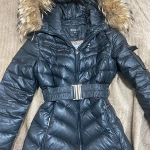 Svart dunjacka med fuskpäls från Hollies - Snygg svart dunjacka från Hollies med avtagbar huva och fuskpäls. Jackan har markerad midja med brett bälte, dragkedja framtill och flera fickor med dragkedja. Quiltad design och lång passform, perfekt för kalla dagar.