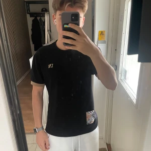 Svart Burberry t-shirt med logga - Svart t-shirt från Burberry i 100% bomull med beige muddar på ärmarna, guldfärgad broderad logga på bröstet och en patch med klassiskt Burberry-mönster vid nederkanten. Snygg och stilren modell med normal passform.
