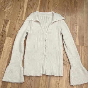 Beige stickad kofta med volangärm - Superfin beige stickad kofta med bred krage och v-ringning. Koftan har långa ärmar med volangdetalj vid ärmsluten och ribbad struktur. Perfekt för lager-på-lager och ger en chill men ändå trendig vibe. Har i storlek M men funkar som S