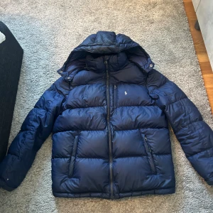 Mörkblå pufferjacka från Polo Ralph Lauren - Säljer en mörkblå pufferjacka från Polo Ralph Lauren med huva och dragkedja framtill. Jackan har två sidofickor med dragkedja och en bröstficka, samt klassisk Polo-logga broderad på bröstet. Perfekt för kalla dagar och riktigt snygg streetstil.