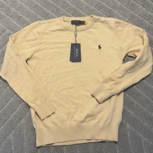 Beige stickad tröja från Polo Ralph Lauren - Beige stickad tröja från Polo Ralph Lauren med klassisk rund halsringning och broderad marinblå logga på bröstet. Tröjan har ribbade muddar vid ärmslut och nederkant, och är långärmad. Perfekt för lager-på-lager och stilren look.