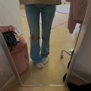 Ljusblå bootcut jeans med hål - Säljer ett par ljusblå jeans med bootcut-modell och slitna hål på ena knät och låret. Jeansen har klassisk femficksdesign och är tillverkade i denim. Perfekta för dig som gillar en avslappnad och trendig look.