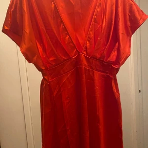 Röd satinklänning med v-ringning toppen  - Supersnygg röd kort klänning i glansig satin med djup v-ringning och markerad midja. Klänningen har korta ärmar och en flowy kjol som ger en riktigt trendig look. Perfekt för dig som vill sticka ut och känna dig extra glam.