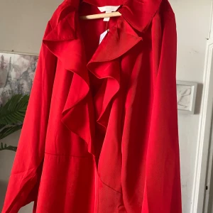 Röd långklänning med volangkrage - Snygg röd långklänning med breda ärmar och en stor volangkrage framtill. Klänningen har en flowy passform och är tillverkad i ett mjukt tyg som faller fint. Perfekt för dig som vill sticka ut med en färgstark look.