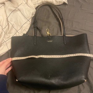 Svart axelväska från Lauren Ralph Lauren - Stilren svart axelväska i skinn från Lauren Ralph Lauren med guldiga detaljer och rejäla handtag. Väskan har en klassisk, rymlig form och subtilt präglad yta. Perfekt för dig som vill ha en snygg och praktisk väska med lyxig känsla.