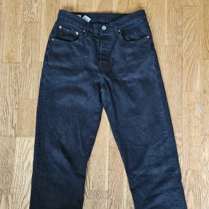 Levi's 501 svarta jeans W26 L28 - Svarta Levi's Premium 501 jeans med klassisk femficksdesign och raka ben. Jeansen har silverfärgade nitar och knapp, samt läderpatch bak i midjan. Tillverkade i slitstark bomullsdenim med en snygg mörk finish. Inga skador men lite tvättblekta. 