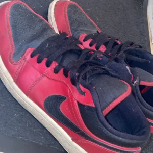 Nike Jordan 1 Low röd och svart - Säljer ett par Nike Jordan 1 Low sneakers i rött och svart med klassisk swoosh på sidan. Skorna har snörning, platt sula och rund tå. Ovandelen är i skinn och textil, vilket ger en snygg kontrast mellan färgerna. Perfekt för dig som gillar streetwear och ikoniska sneakers.