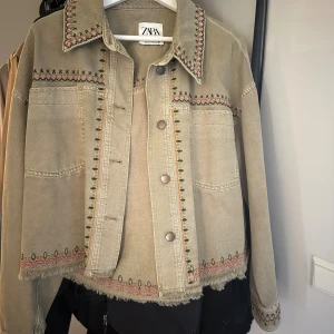 Beige broderad jeansjacka från Zara - Snygg beige/khaki jeansjacka från Zara med broderade detaljer i rosa, grönt och svart på krage, axlar, ärmslut och nederkant. Perfekt för dig som gillar unika detaljer och chill stil. Passar även xs