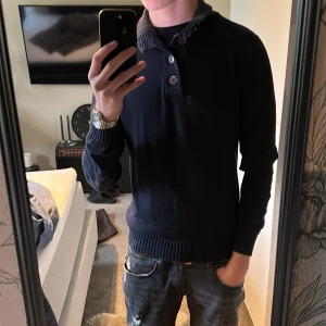 Mörkblå stickad tröja med knappar - Mörkblå stickad tröja med ribbad krage och tre knappar framtill. Tröjan har lång ärm och en lite loose passform, perfekt för kyligare dagar. Snygg och enkel att matcha med jeans eller andra byxor.