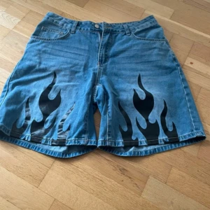 Blå jeansshorts med flames - Säljer ett par blå denimshorts med svarta flames tryckta framtill. Klassisk femficksmodell med knapp och dragkedja. Shortsen har en loose fit och är perfekta för dig som vill sticka ut med en cool detalj. Passar till sommaren och har en avslappnad vibe.