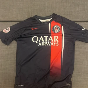 PSG Mbappé 7 fotbollströja Nike - Paris Saint-Germain matchtröja med Mbappé 7 på ryggen. Tröjan är mörkblå med en röd vertikal rand och vita detaljer, PSG-emblem på bröstet och Nike-logga i rött. Tillverkad i lätt polyester med Dri-FIT ADV-teknologi. Snygg design för dig som älskar fotboll och PSG.