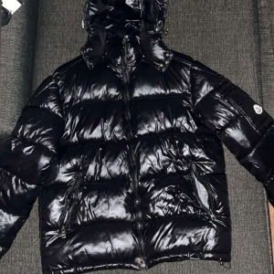 Svart Moncler pufferjacka med huva - Säljer en svart pufferjacka från Moncler med glansig finish och huva. Jackan har dragkedja framtill, två sidofickor med dragkedja och en ficka med Moncler-logga på ärmen. Insidan har ett coolt serietryck. Perfekt för dig som vill ha en snygg och varm jacka.