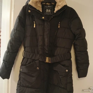 Svart pufferjacka med bälte Havana - Svart pufferjacka från Hava by Havana med snyggt skärp med guldspänne i midjan . Jackan har långa ärmar, flera fickor med knappar och dragkedjor i guld. Perfekt för kalla dagar med sin quiltade design och varma material. Fuskpäls fattas på luvan samt en knapp på ena fickan.