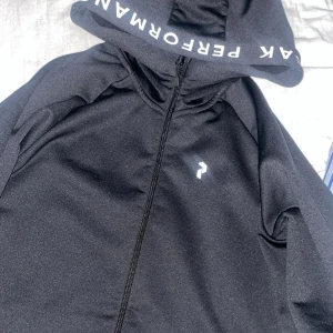Peak performance zip hoodie med text på luvan - Svart kofta från Peak Performance med dragkedja framtill och diskret logga på bröstet. Jackan har huva och praktiska fickor med dragkedja. Perfekt för dig som gillar en sportig och clean stil. Den är storlek 170 vilket motsvarar Xs köpt från kidsbrandstore för 1200 helt oanvänd