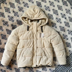 Beige pufferjacka från IETS FRANS - Beige pufferjacka från IETS FRANS med broderad logga fram och bak. Jackan har huva med svarta dragsnören, två snedställda fickor och dragkedja framtill. Ytan är mjuk fleece och jackan är quiltad för extra värme. Perfekt för kalla dagar. 🤎
