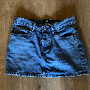 Blå jeanskjol från 157, XS - Snygg blå jeanskjol från 157 i klassisk denim med fem fickor och bälteshällor. Kjolen har en rak passform och stängs med knapp och dragkedja framtill. Perfekt för dig som gillar en enkel och tidlös stil.  PRIS KAN DISKUTERAS