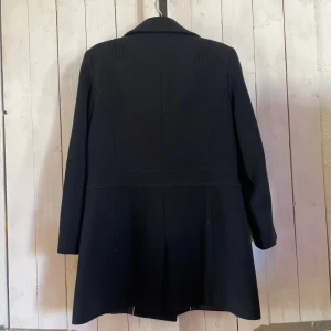 Svart kappa från Quiz, UK 12 - Stilren svart kappa från Quiz med dubbelknäppning och bred krage. Jackan har dekorativa knappar framtill, två fickor med lock och markerad midja. Perfekt för dig som vill ha en klassisk och snygg look under höst och vinter.