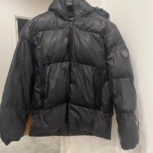Svart pufferjacka från EA7 Emporio Armani - Säljer en svart pufferjacka från EA7 Emporio Armani med huva och dragkedja. Jackan har en stilren look med EA7-logga på ärmen och två fickor framtill. Perfekt för kalla dagar när du vill hålla stilen och värmen.