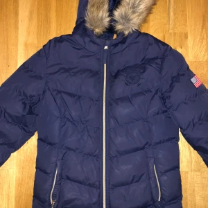 Blå pufferjacka Vinson Polo Club S - Blå pufferjacka från Vinson Polo Club med huva och fuskpäls. Jackan har dragkedja framtill, två fickor med dragkedja och amerikansk flagga på ärmen. Broderad logga på bröstet och quiltad design. Perfekt för kalla dagar.Skick: Jackan är i princip som ny! Jag har bara använt den ett par gringar, så den är i felfritt skick.