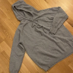 Grå hoodie med snörning - Mysig grå kashmir  hoodie med huva och snörning framtill. Tröjan har långa ärmar och ribbade muddar vid ärmslut och nederkant. Perfekt för chill dagar eller när du vill ha en enkel och stilren look.