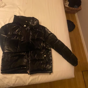 Svart pufferjacka från Stradivarius - Säljer en svart, glansig pufferjacka från Stradivarius. Jackan har hög krage, dragkedja framtill och tryckknappar vid fickorna. Tillverkad i syntetmaterial och designad för att stå emot både vind och regn. Perfekt för kalla dagar och har en riktigt modern vibe.