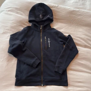 Mörkblå hoodie från Sail Racing - Mörkblå hoodie från Sail Racing med dragkedja framtill och bröstficka med dragkedja. Broderad logga på huvan och bröstet. Skön passform och tillverkad i mjuk bomull. Perfekt för kyliga dagar och snygg till jeans.