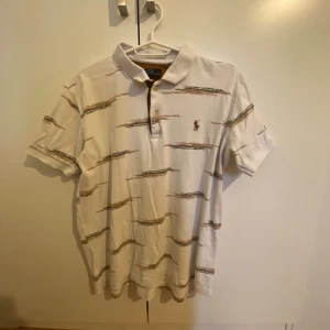 Vit randig piké från Polo Ralph Lauren - Vit pikétröja från Polo Ralph Lauren med bruna och svarta horisontella ränder och klassisk broderad logga på bröstet. Skjortan har krage, knappar framtill och korta ärmar. Perfekt för en chill och stilren look.