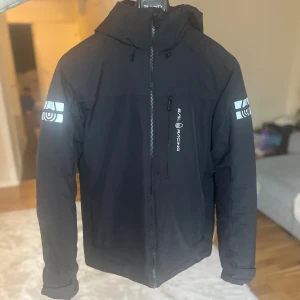 Svart Sail Racing jacka med huva - Säljer en svart jacka från Sail Racing med huva och dragkedja framtill. Jackan har logga på bröstet och tryck på båda ärmarna. Den är långärmad och har en sportig look, perfekt för dig som gillar stilrena och funktionella plagg. Storlek M