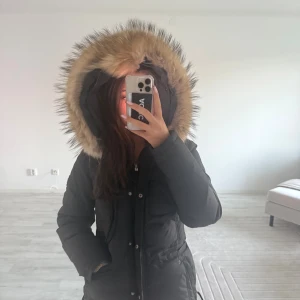 Svart parkas med äkta päls från Hollies - Säljer en svart parkas från Hollies med stor huva kantad av beige fuskpäls. Jackan har dragkedja och knappar framtill, samt praktiska fickor och en figurnära passform. Perfekt för kalla dagar och riktigt snygg till vintern.